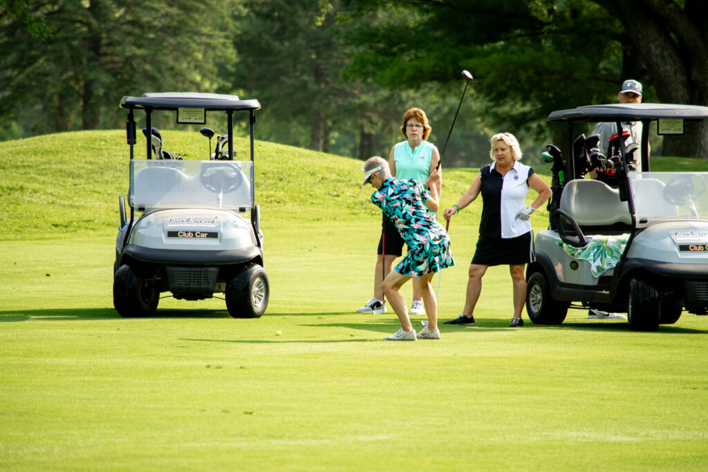 group of golfers watching a woman take a swing on the course near two golf carts